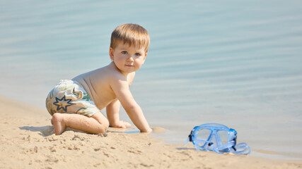Toddler crawling on sandy shore near blue water with curious expression. Concept of childhood, discovery, play, leisure, innocence, summer holiday and family lifestyle.