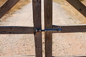 Close-up of a wooden fence gate with metal bolt and padlock in a rural field in Spain. Textured gravel road emphasize traditional farmland boundaries and the character of southern Spain’s landscape