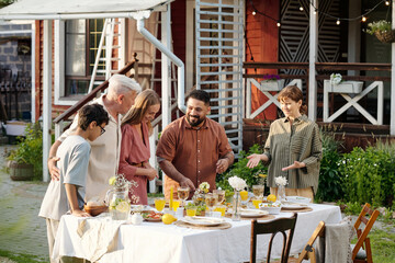 Multiethnic group including senior Caucasian man, middle aged Caucasian woman, young adult couple, teenage boy gathering around outdoor table sharing meal and conversation