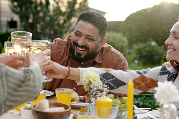 Middle Eastern man smiling while clinking glasses with diverse group of adults outdoors, enjoying meal together at table with food and drinks, greenery in background