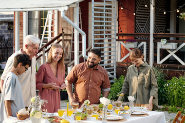 Group of multiethnic adults and one teenager gathering around outdoor table sharing meal and smiling, standing in backyard setting with food and drinks arranged on table