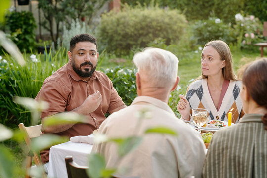 Middle Eastern man talking while sitting at outdoor table with middle aged Caucasian woman and senior Caucasian man, group sharing meal in garden setting, people interacting