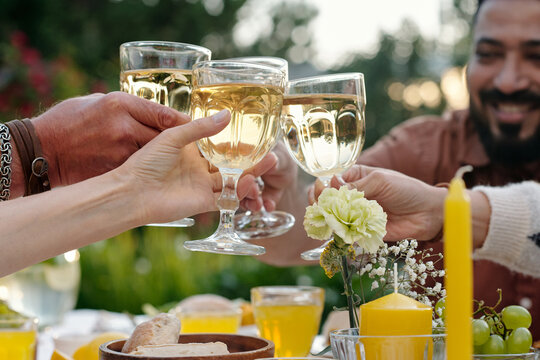 Group of multiethnic friends clinking wine glasses during outdoor gathering, hands visible holding drinks above table with food and candles, smiling man in background