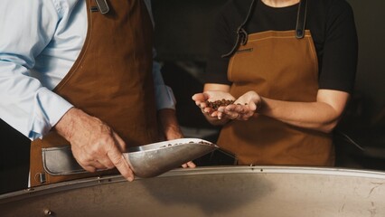 Two workers in aprons standing in coffee factory. One holding metal scoop with roasted beans while other dropping them from his hands. Examining quality and discussing roasting results.