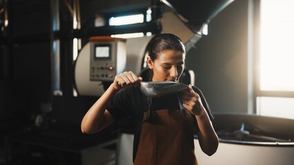 Caucasian woman in brown apron holding metal scoop close to face and smelling freshly roasted...