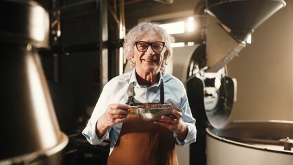 Smiling man in brown apron and blue shirt holding metal bowl of roasted coffee beans. Looking directly at camera in coffee roasting facility with industrial roasting machine in background.