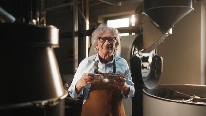 Smiling man in brown apron and blue shirt holding metal bowl of roasted coffee beans. Looking directly at camera in coffee roasting facility with industrial roasting machine in background.