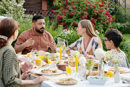 Multiethnic group of middle aged man, Caucasian woman, senior woman, young boy and others sitting outdoors around table sharing meal and talking during garden gathering