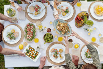 Group of multiethnic adults and teenager sharing food around outdoor table, passing plates and reaching for dishes, enjoying meal together, hands visible from overhead perspective