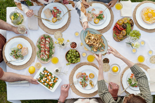 Group of multiethnic adults and teenager sharing food around outdoor table, passing plates and reaching for dishes, enjoying meal together, hands visible from overhead perspective - Powered by Adobe