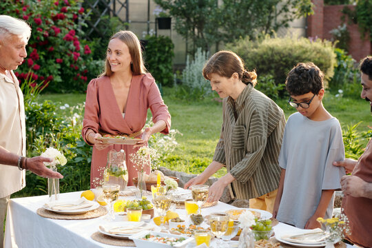 Caucasian young adult woman serving food to senior Caucasian man, while middle aged Caucasian woman arranging table and teenage boy with glasses standing beside multiethnic group outdoors