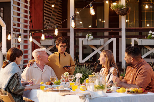 Multiethnic group of middle aged and young adult men and women sitting at outdoor table eating dinner, while teenage boy standing holding smartphone and talking to group - Powered by Adobe