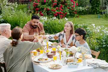 Multiethnic group of grandparents, young married couple and their child toasting drinks while sitting at outdoor table sharing meal