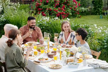Multiethnic family including young parents, senior grandparents and boy sitting outdoors enjoying meal together at garden table, smiling and talking