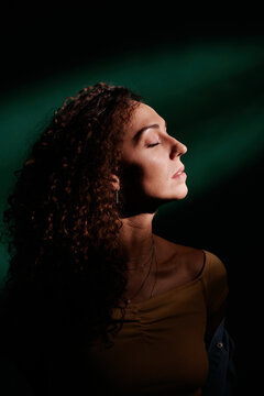 Studio portrait shot of attractive young adult Caucasian woman with curly hair closing her eyes standing in dark room with light hitting her face
