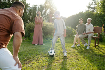 Teenage boy playing soccer with adult man outdoors while Caucasian woman taking photo on smartphone and senior Caucasian couple sitting on bench watching game