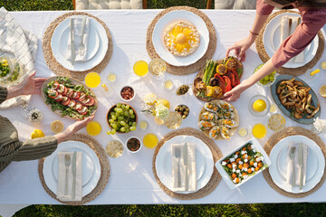 Three Caucasian women arranging assorted vegetable dishes and appetizers on outdoor dining table set for meal, hands visible reaching for plates, fresh fruit and juice on table