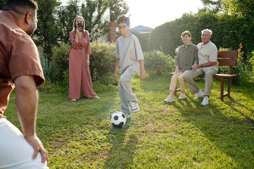 Teenage boy playing soccer with adult man outdoors while middle aged Caucasian woman photographing scene and senior couple sitting nearby