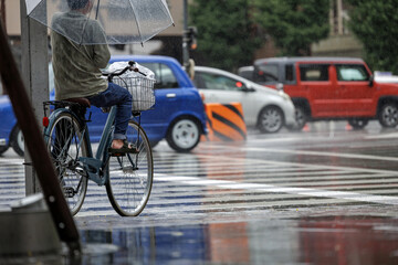 Person on bicycle shelters under clear umbrella while waiting at busy intersection crosswalk. Dressed casually in jeans, sandals, and waffle shirt, local resident goes shopping during a rainfall.