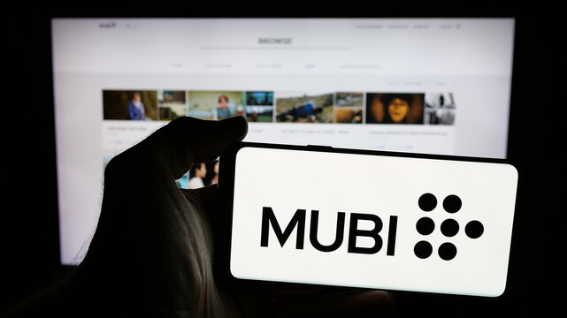 Stuttgart, Germany - 08-20-2025: Person holding mobile phone with logo of British streaming platform company Mubi on screen in front of web page. Focus on phone display.