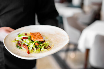 Close-up of a person holding a plate with grilled salmon on mixed vegetable salad, served in a restaurant setting. Healthy gourmet meal presentation.