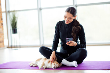 Charming woman sitting on yoga mat with smartphone, petting her dog, enjoying a moment of relaxation in a modern indoor setting