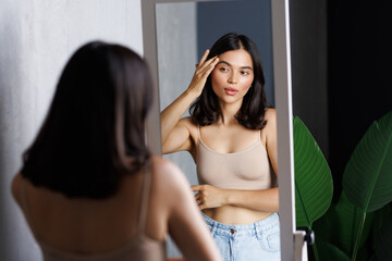 Fototapeta premium Happy beautiful young lady touching face look in bathroom mirror
