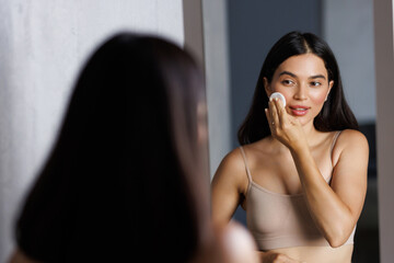 Smiling young woman wiping her face with cotton pad in the bathroom. Skin care concept