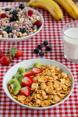 Bowl of cornflakes with fresh strawberries and kiwi on red checkered tablecloth, served alongside colorful fruit cereal, blueberries, bananas, strawberries, and a glass of milk