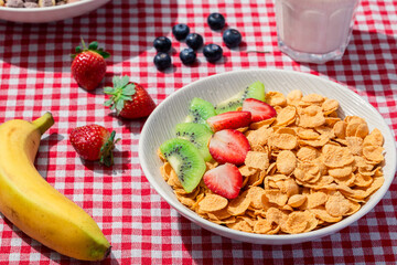 Bowl of cornflakes with strawberries and kiwi on red checkered tablecloth, served with banana, blueberries, colorful cereal, fresh strawberries, and a glass of milk for breakfast