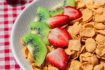 Close up of cornflakes served with fresh sliced strawberries and kiwi on red checkered tablecloth, showing healthy breakfast concept with vibrant colors, texture, and natural detail