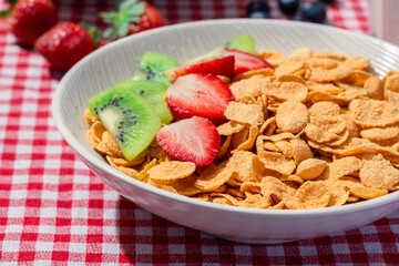Bowl of cornflakes with strawberries and kiwi on red checkered tablecloth, served with banana, blueberries, colorful cereal, fresh strawberries, and a glass of milk for breakfast