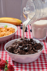 Milk pouring into bowl of chocolate cereal on red checkered tablecloth, with cornflakes, banana, strawberries, and blueberries in background, showing sweet breakfast and lifestyle nutrition