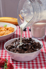Milk pouring into bowl of chocolate cereal on red checkered tablecloth, with cornflakes, banana, strawberries, and blueberries in background, showing sweet breakfast and lifestyle nutrition