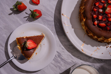 Top view of baked cheesecake with strawberry topping on white plate, served with a slice on side plate, fresh strawberries, and glass of milk on marble background