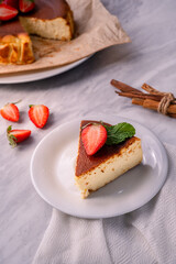 Slice of baked cheesecake topped with strawberry and mint on white plate, with whole cake and cinnamon sticks in background, showing creamy dessert and rustic homemade presentation