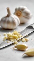 Chopped garlic cloves sit on a stainless steel knife beside whole bulbs in the kitchen