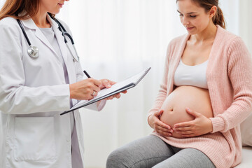 Caucasian pregnant young adult woman sitting on examination table holding belly while female doctor writing on clipboard during prenatal checkup in medical office