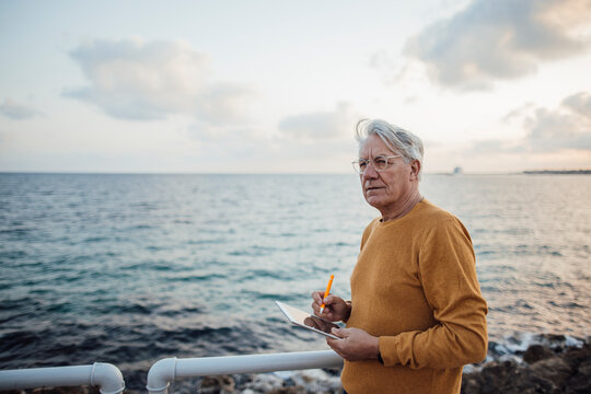 Thoughtful senior man standing with tablet PC in front of sea at sunset