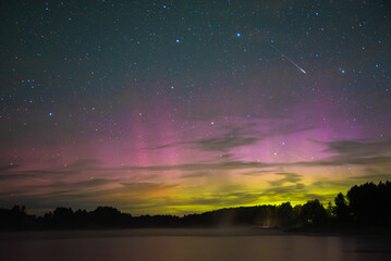 Fototapeta premium Colorful aurora borealis in green and pink shades over a calm lake with forest silhouettes and a shooting star in the starry night sky.