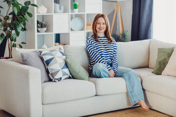 Obraz premium Happy woman in a stylish striped top enjoying a comfortable and cozy moment on a chic sofa in a modern living room