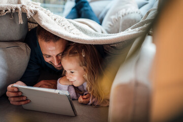 Happy man sharing tablet computer with daughter under blanket at home