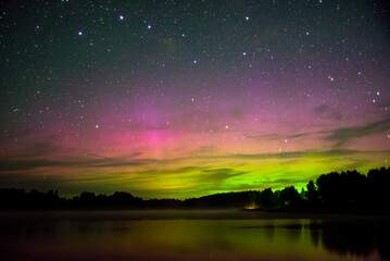 Naklejka premium Beautiful aurora borealis in green and pink hues reflecting over a calm lake under a starry night sky with forest silhouette.