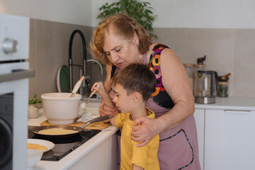 A cute, beautiful, happy Grandma is teaching her little grandson how to fry pancakes in a frying pan. The boy is happy and smiling. Family values. A beloved grandson. Cooking together.