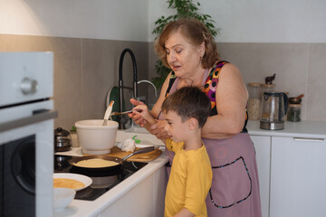 A cute, beautiful, happy Grandma is teaching her little grandson how to fry pancakes in a frying pan. The boy is happy and smiling. Family values. A beloved grandson. Cooking together.