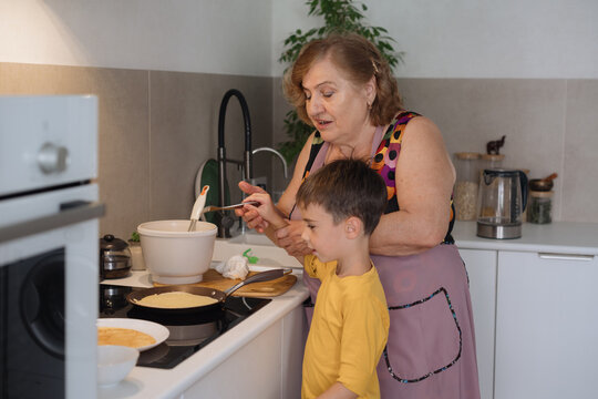 A cute, beautiful, happy Grandma is teaching her little grandson how to fry pancakes in a frying pan. The boy is happy and smiling. Family values. A beloved grandson. Cooking together.