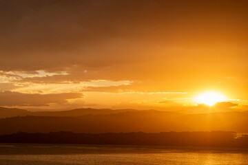Obraz premium Fiery sunset at Merimbula, Sapphire Coast New South Wales, Australia. Orange sky glowing beneath clouds across coastal hills, calm ocean and lake, creating a dramatic, tranquil seascape at dusk.
