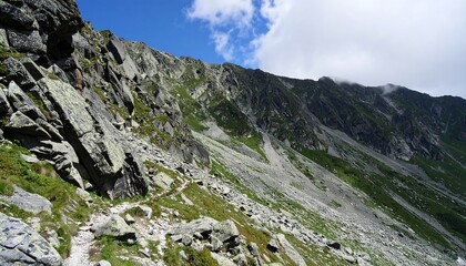 A rugged mountain trail winds through a landscape of large rocks and greenery under a partly cloudy sky.