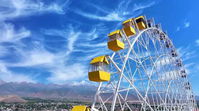 Giant ferris wheel against the background of vibrant blue sky and high mountains.  4K UHD drone video of yellow observation wheel on a sunny day. Attraction, vacation, holidays, rest concept