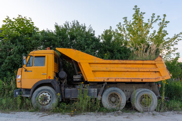 Obraz premium Old yellow dump truck parked in overgrown area with wild grass and trees under clear sky. Concept of abandoned industrial equipment, rural decay and nature reclaiming urban infrastructure.